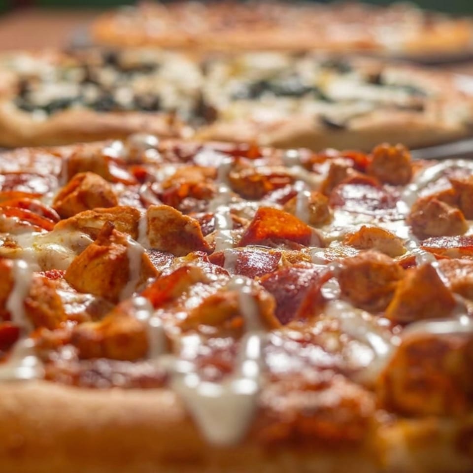 Close-up of a loaded pizza topped with chicken, pepperoni, vegetables, melted cheese, and creamy drizzle, with another pizza in the background.