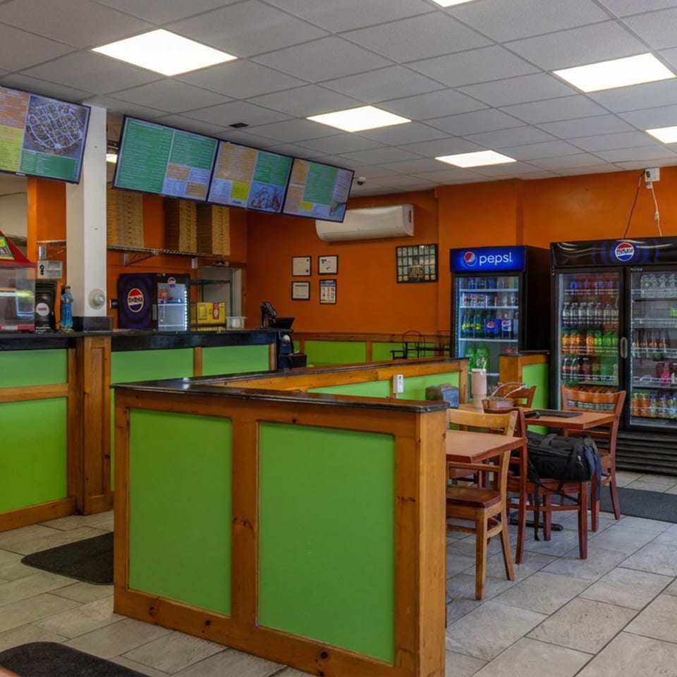 Bright casual restaurant interior with green-and-wood counters, menu screens, beverage refrigerators, kitchen equipment, and tables.